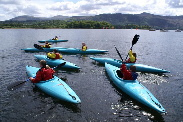  6 niños con chalecos salvavidas rojos y amarillos haciendo kayak en el lago 