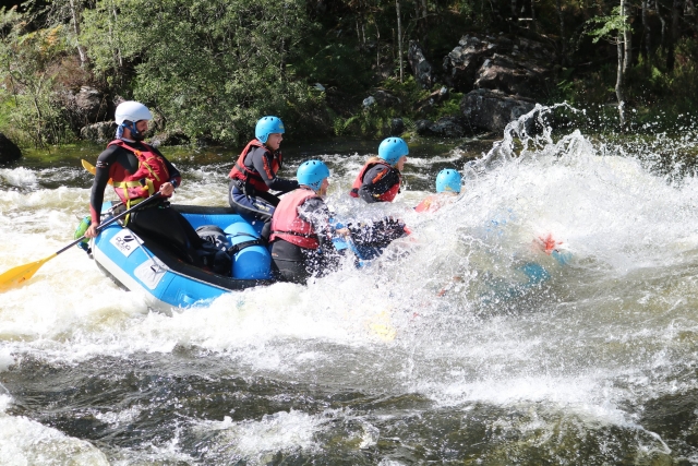  Personas con chalecos salvavidas rojos y cascos azules golpeados por una ola en el río mientras practican rafting
