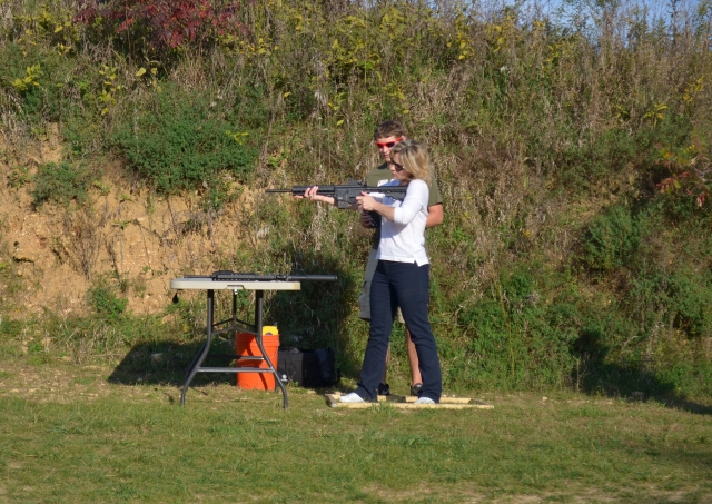  Mujer rubia con chamarra blanca disparando con rifle 