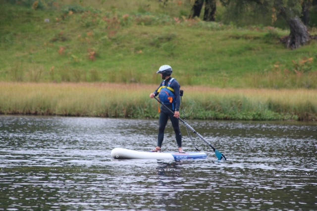  Chico con traje de neopreno azul y chalecos de flotación azules haciendo paddleboarding y bosque al fondo 