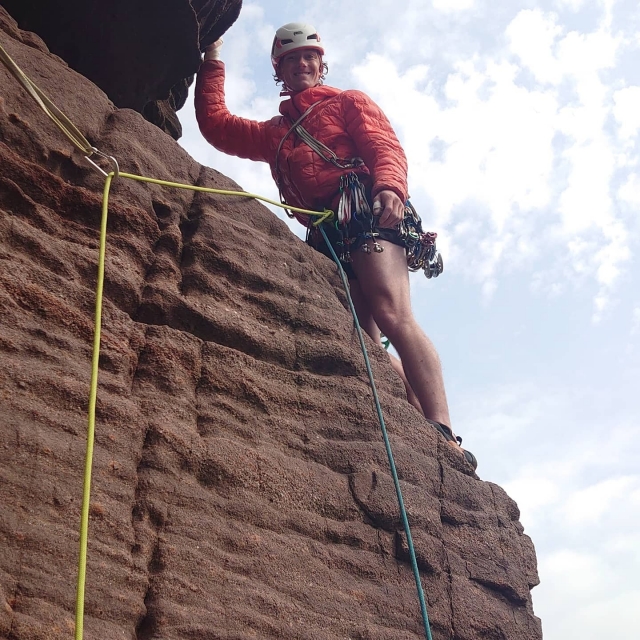  Hombre con chamarra naranja, pantalón azul, casco blanco, sonriendo y escalando 
