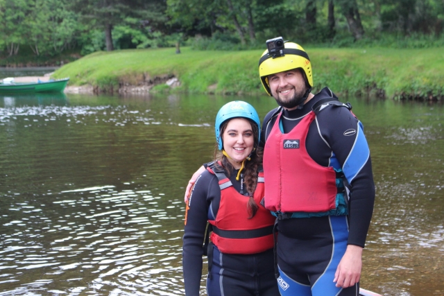 Pareja sonriendo y vistiendo chalecos salvavidas rojos y cascos azules y amarillos a punto de disfrutar de su sesión de piragüismo