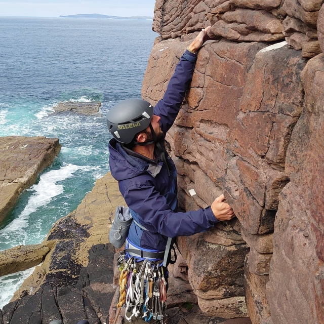 Niño con casco negro y chamarra azul escalando y el mar al fondo