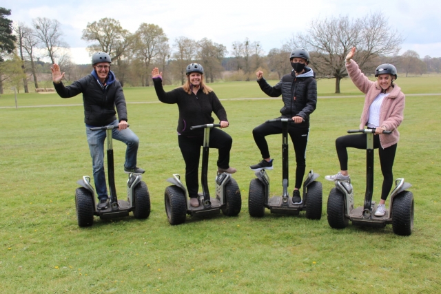  Cuatro amigos con cascos negros levantando la mano y sonriendo mientras hacen segway en el parque 