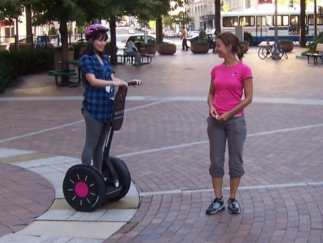  Chica con casco rosa, sonriendo y recibiendo información de su instructor, que lleva una playera rosa. Ella se prepara para hacer una sesión de segway 
