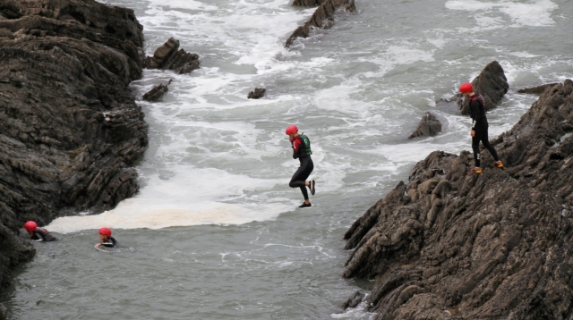 Cuatro personas con trajes de neopreno negros y cascos rojos saltando desde un acantilado 