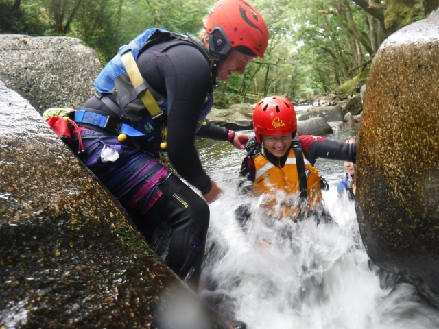 Instructor con casco rojo y ayudas de flotabilidad azules ayudando a un niño usando casco rojo y ayudas de flotabilidad amarillas mientras practica barranquismo