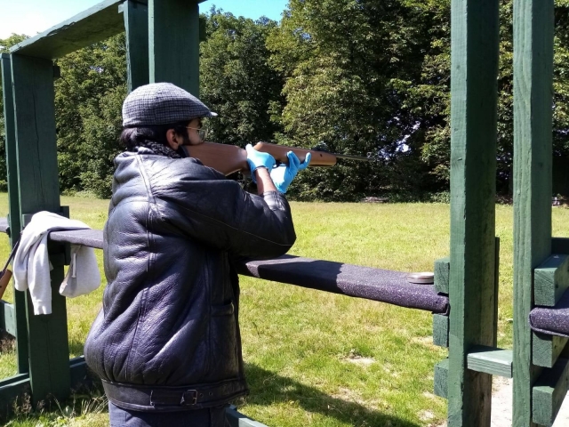 Hombre con chamarra de cuero y gorra disparando con rifle 
