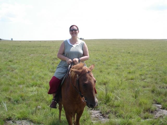  Mujer sonriendo con playera gris subiendo un caballo marrón 
