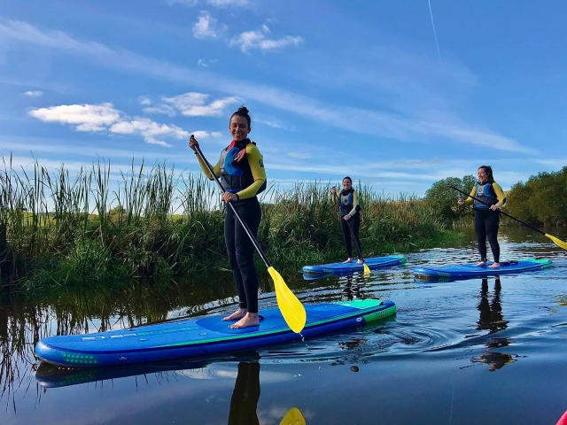  Tres niñas vestidas con trajes de neopreno amarillos y negros y ayudas de flotabilidad negras haciendo paddleboarding 