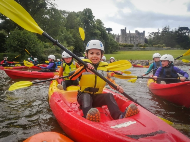  Niños usando ayudas de flotabilidad amarillas navegando en kayak y el castillo de Castlewellan al fondo 