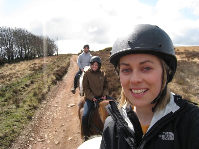 Chica sonriendo con casco negro y suéter con dos amigas a su espalda subiendo a caballo 