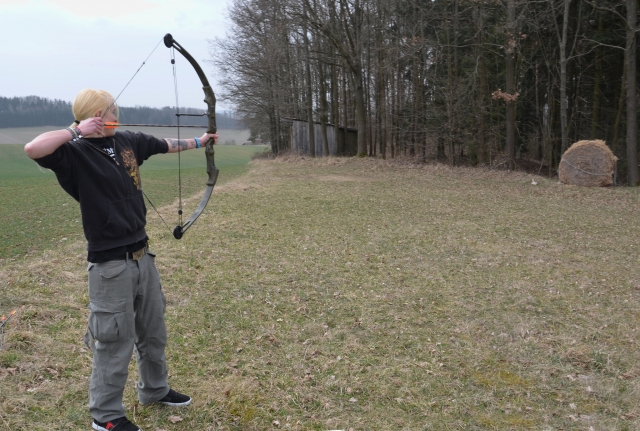 Hombre con cabello largo , vistiendo playera negra y jeans grises, practicando jalo con arco.
