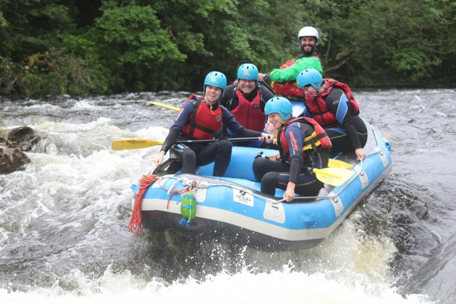 Familia con dos chavos con cascos azules y ayudas de flotabilidad rojas mientras hacen rafting 