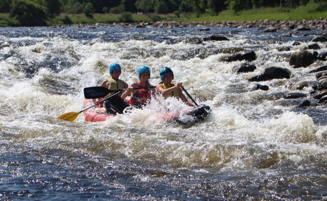 Niñas riendo y usando cascos azules y ayudas de flotabilidad verdes y rojas golpeadas por olas mientras practican rafting 