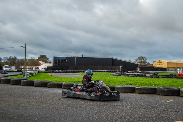  Niña con casco azul y traje de manejar negro karting e instalaciones en la parle trasera 