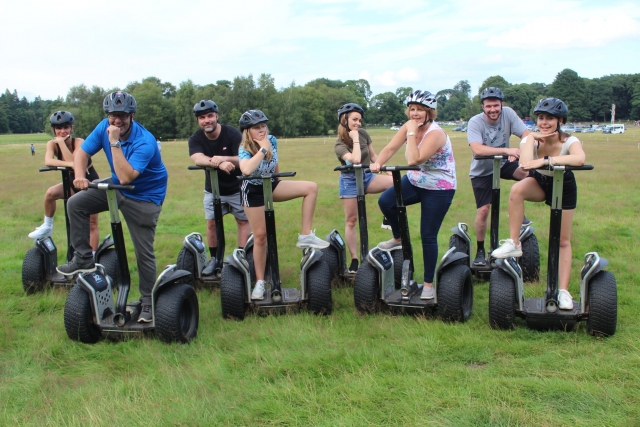  Grupo de niños y niñas con camisetas, cascos grises, sonriendo y paseando en segway por el parque