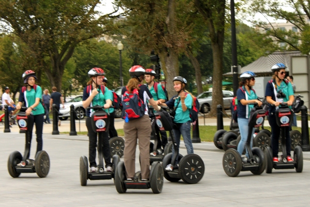  Personas con camisetas azules y cascos rojos y blancos subiendo en segways en el parque 