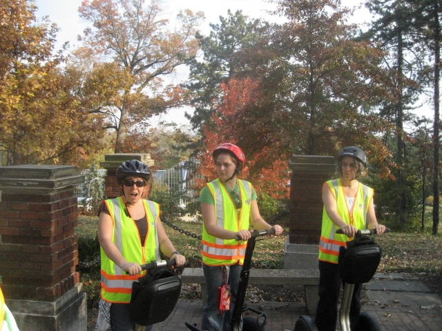  Tres mujes con cascos azules, negros y rojos sonriendo y haciendo un recorrido en segway por el parque 