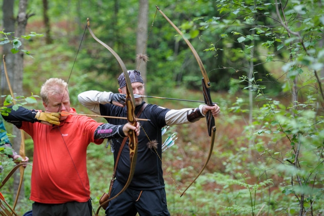 Dos hombres practicando jalo con arco en el bosque, uno con playera roja y el otro con playera azul.