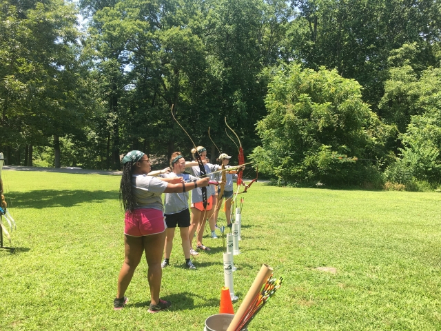  Grupo de chicas practicando jalo con arco debajo la arena y rodeado por el bosque