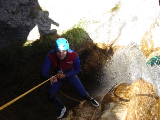  Hombre escalando el acantilado con ayudas de flotabilidad rojas y casco azul duranle su sesión de barranquismo 