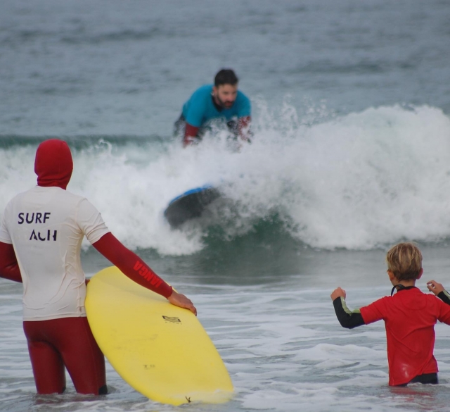 Un surfista de nuestra escuela anle la atenta mirada de un instructor y un niño pequeño